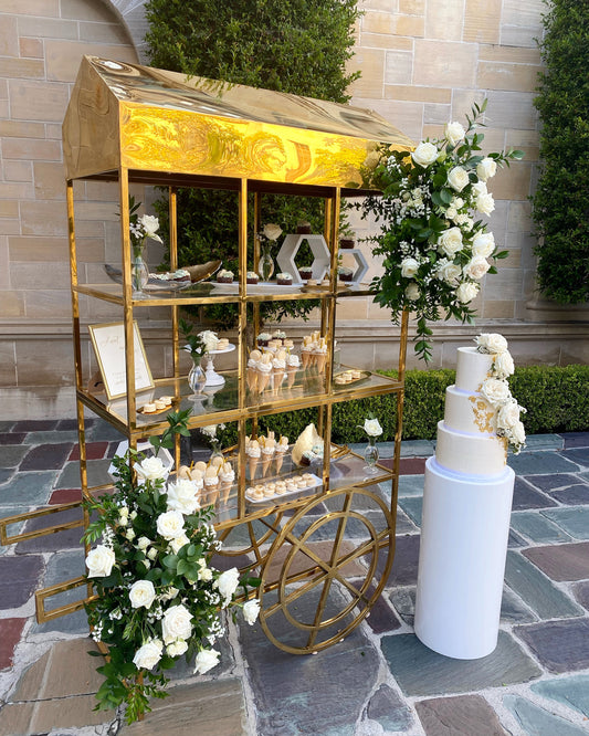 Gold bar cart with wedding cake and flowers on a stone patio