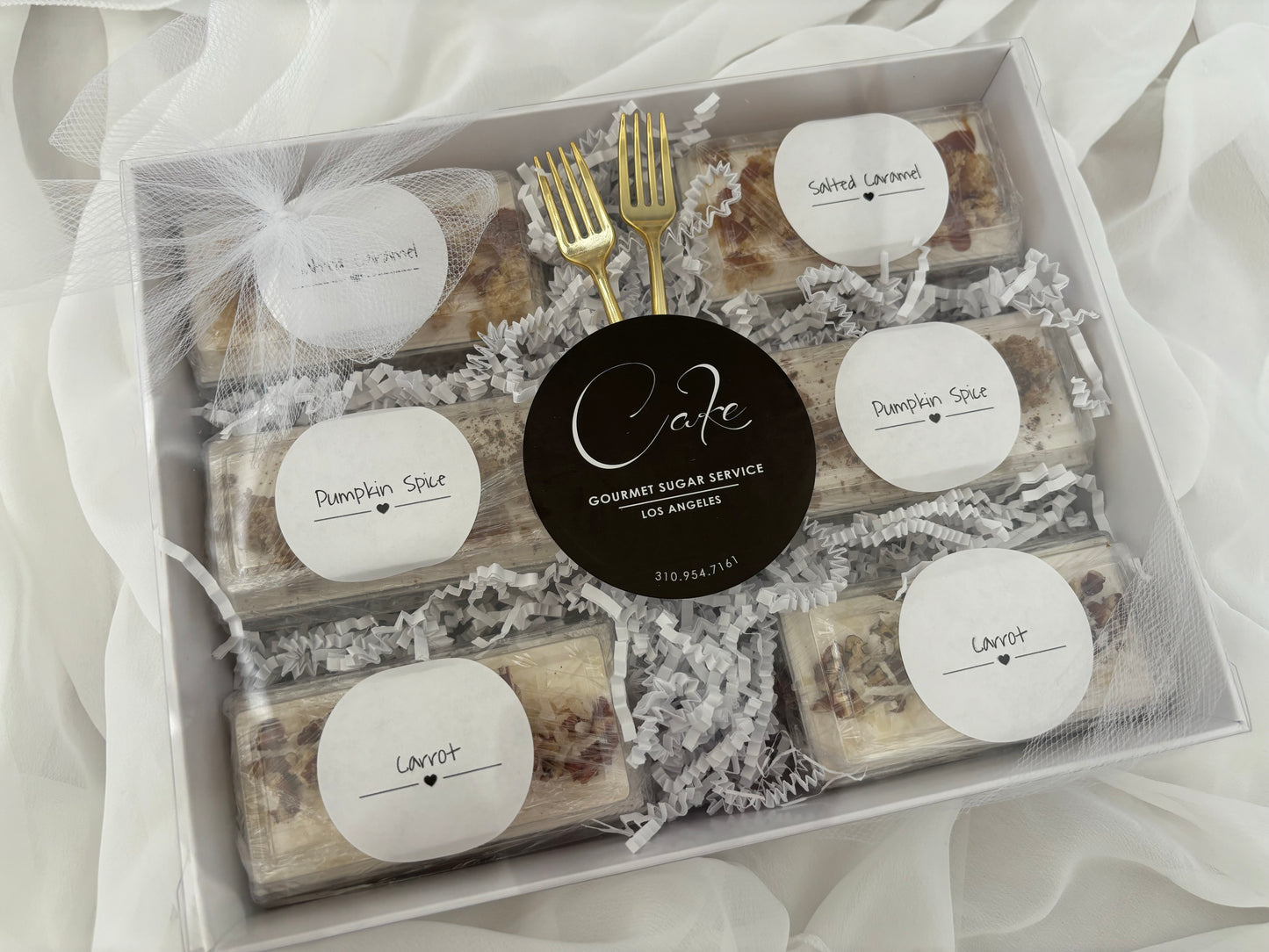 Box of assorted gourmet sugar cookies with labels and a gold fork on a white fabric background.