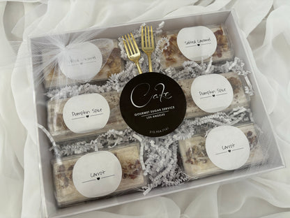 Box of assorted gourmet sugar cookies with labels and a gold fork on a white fabric background.