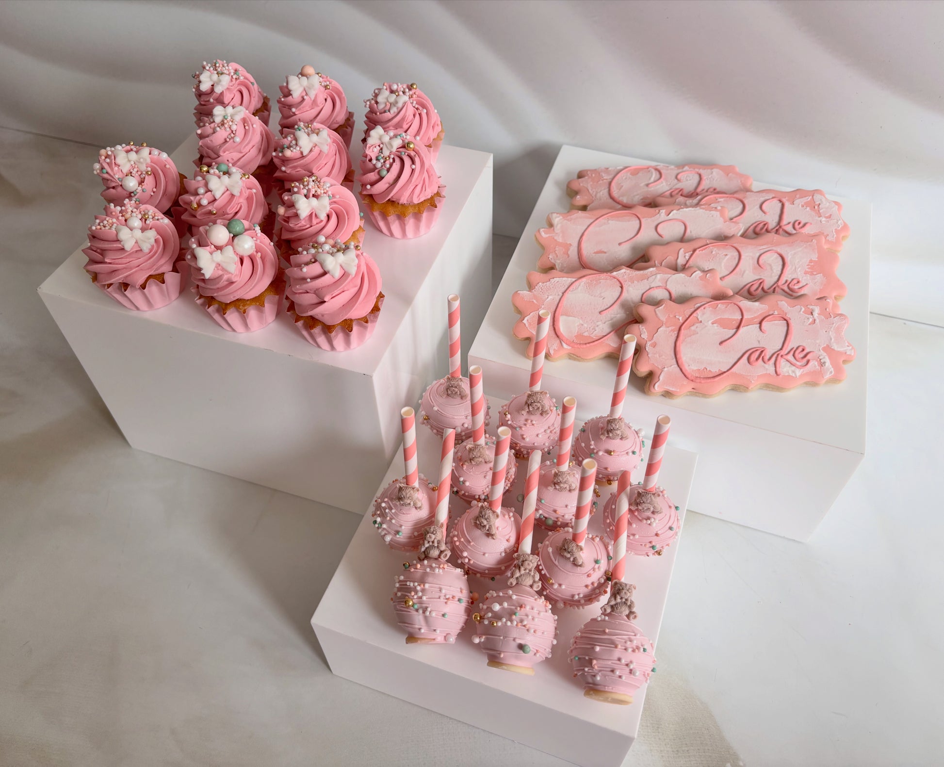 Pink-themed dessert set including cupcakes, cookies, and cake pops on a white surface.