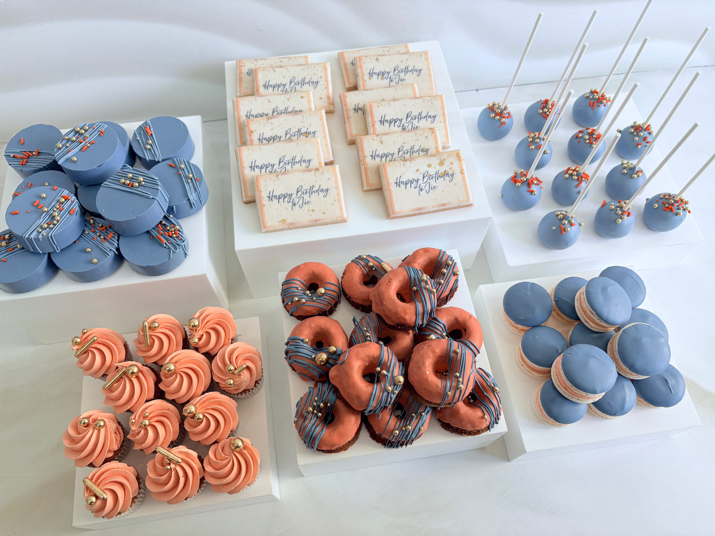 Assorted birthday cakes including cupcakes, donuts, and cookies on a white surface.