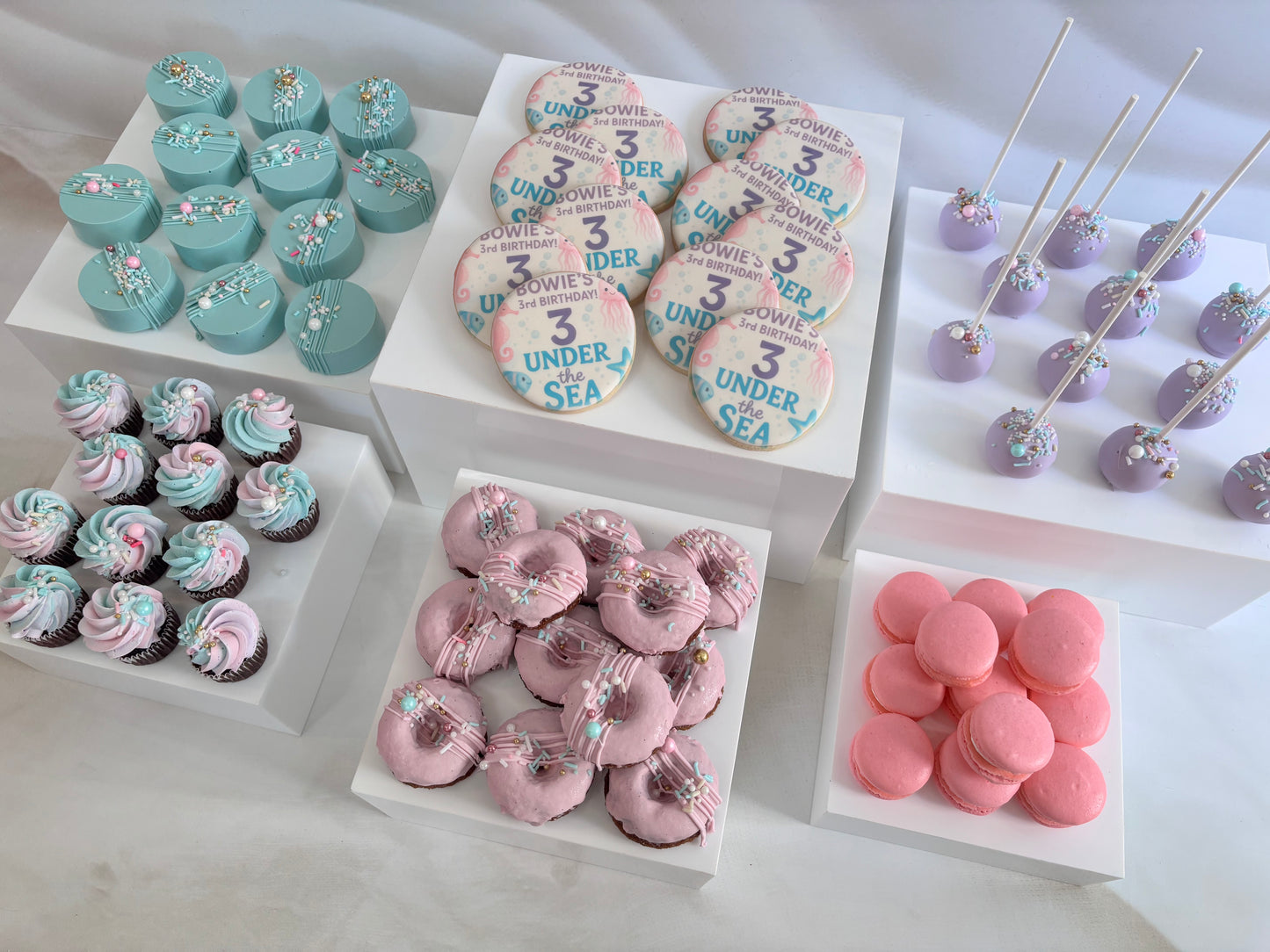 Assorted pastel-colored desserts including cupcakes, cookies, and macarons on a white surface.