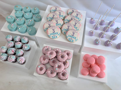 Assorted pastel-colored desserts including cupcakes, cookies, and macarons on a white surface.