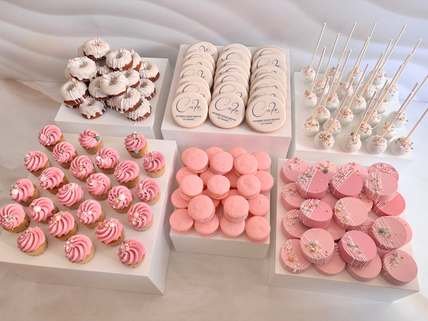 Assorted pink and white desserts including cupcakes, cookies, and macarons on a light background.