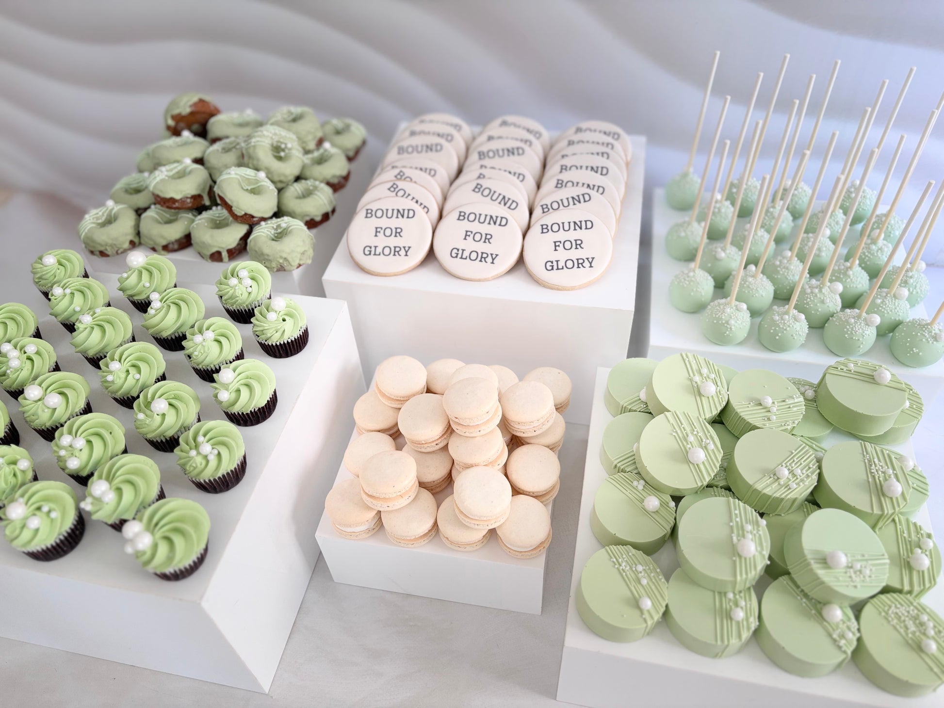 Assorted green and white cupcakes, cookies, and macarons on a light gray background.